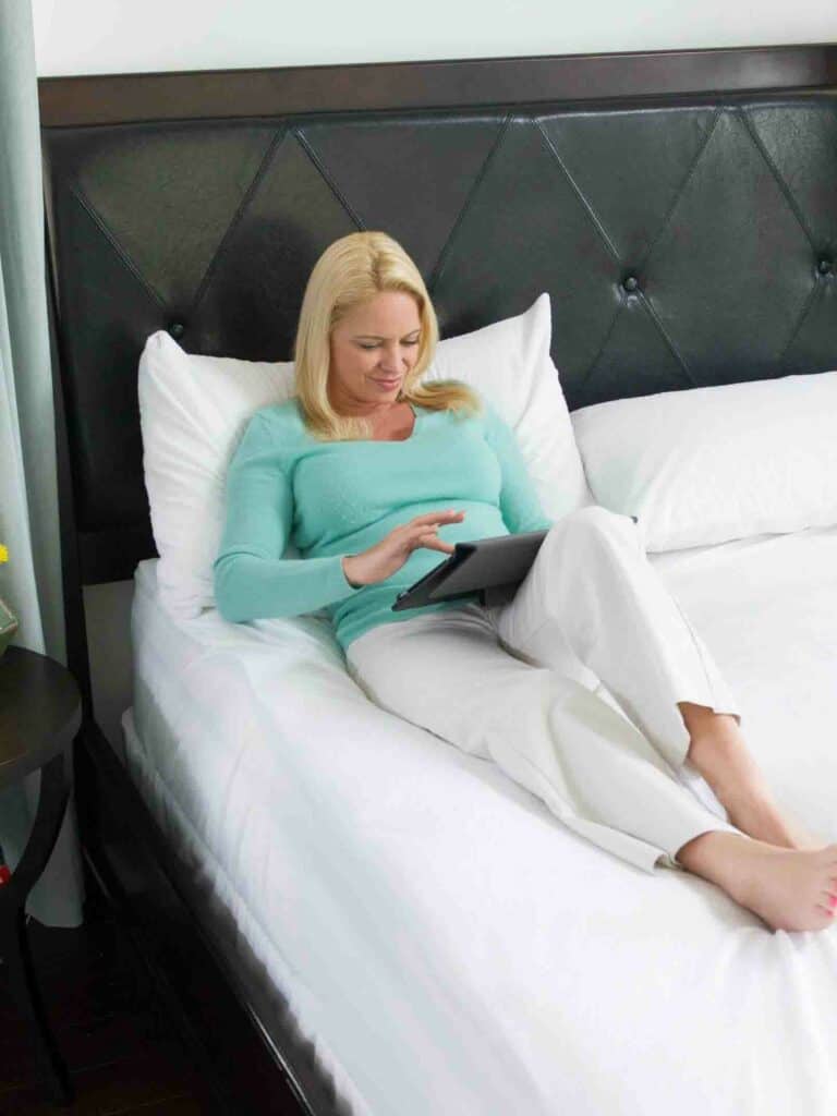 Woman in turquoise top relaxing and reading on a tablet in bed with plush white bedding